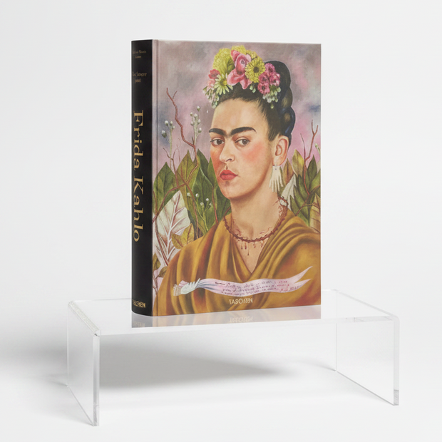 A hardcover art book titled 'Frida Kahlo' by Taschen, featuring a colorful painted portrait of Frida Kahlo on the cover and a dark spine, displayed on a purple surface against a black background.